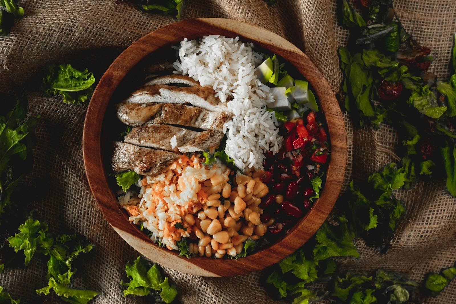 A wooden bowl of sliced spiced chicken with rice, beans and salad