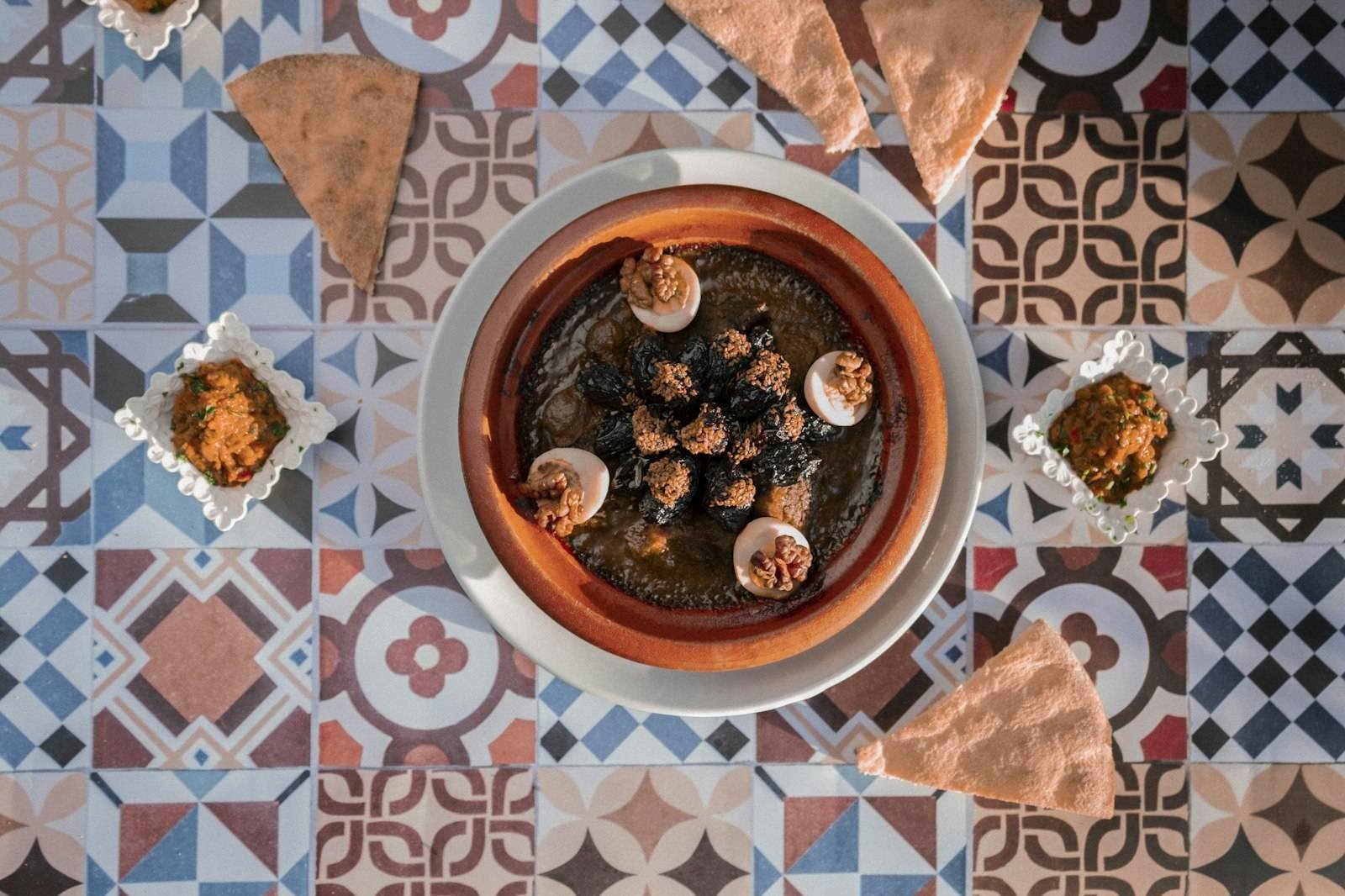 A Moroccan tagine dish viewed from above on a patterned tile table with flatbread